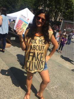 long haired person wearing a gold vest reading, “Ask me about fake clinics!” and holding informational pamphlets on fake clinics
