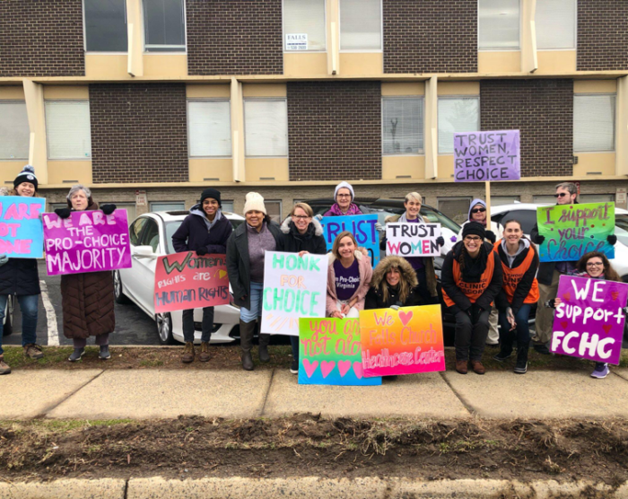 Image: A group of 14 people, including activists and clinic escorts, outside of the Falls Church Healthcare Center with signs in support of the clinic with messages such as, “You are not alone” and “Trust women”
