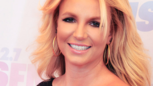 Image Description: Britney Spears, a white woman with bright blond hair, is smiling at the camera. She is wearing gold hoops and a black dress with one sleeve.]