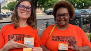 Picture of Reproaction co-founders and co-directors Erin Matson and Pamela Merritt wearing orange shirts with text on the front: Stop Prosecuting Abortion