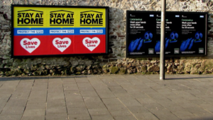 A picture of a worn, brown stone wall in Newport City Centre. There are two sets of three billboards on the wall: One set is yellow, blue, and red and says “Stay at home, protect the NHS, save lives”; the other is black and blue with white text that discusses coronavirus hand washing protocols.